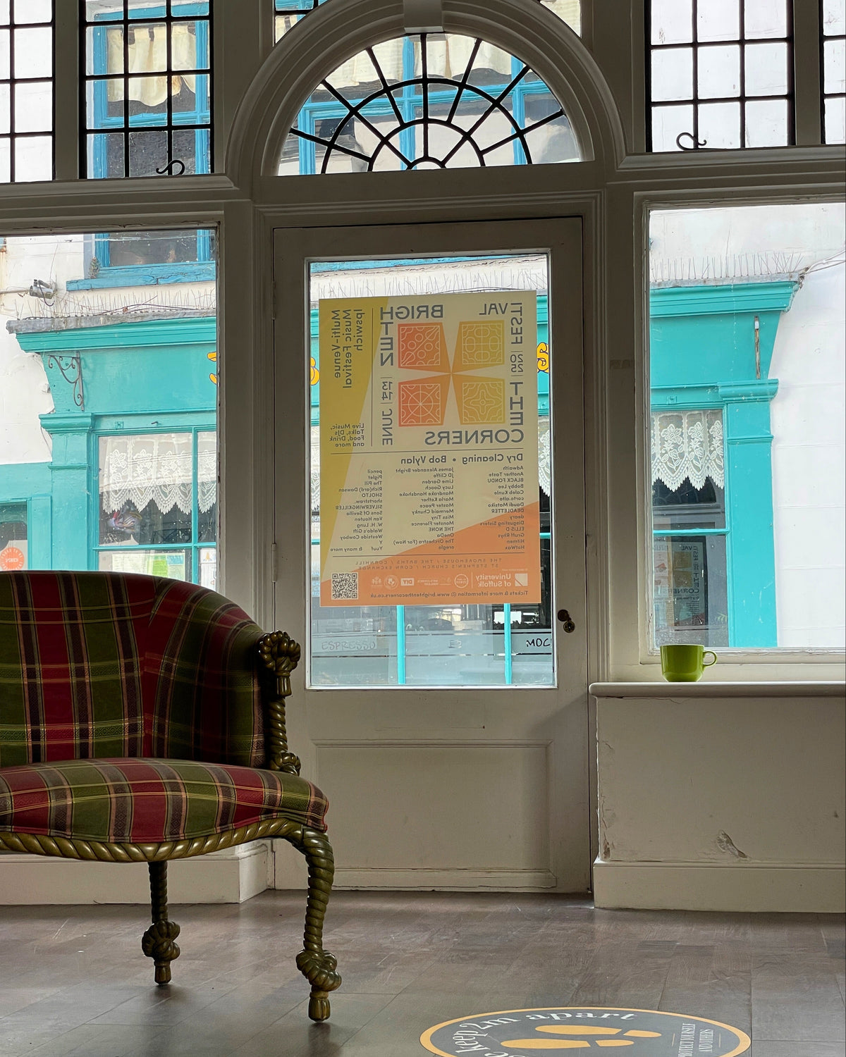 Plaid armchair in a room with large windows showing a white building exterior and Brighten The Corners festival poster.